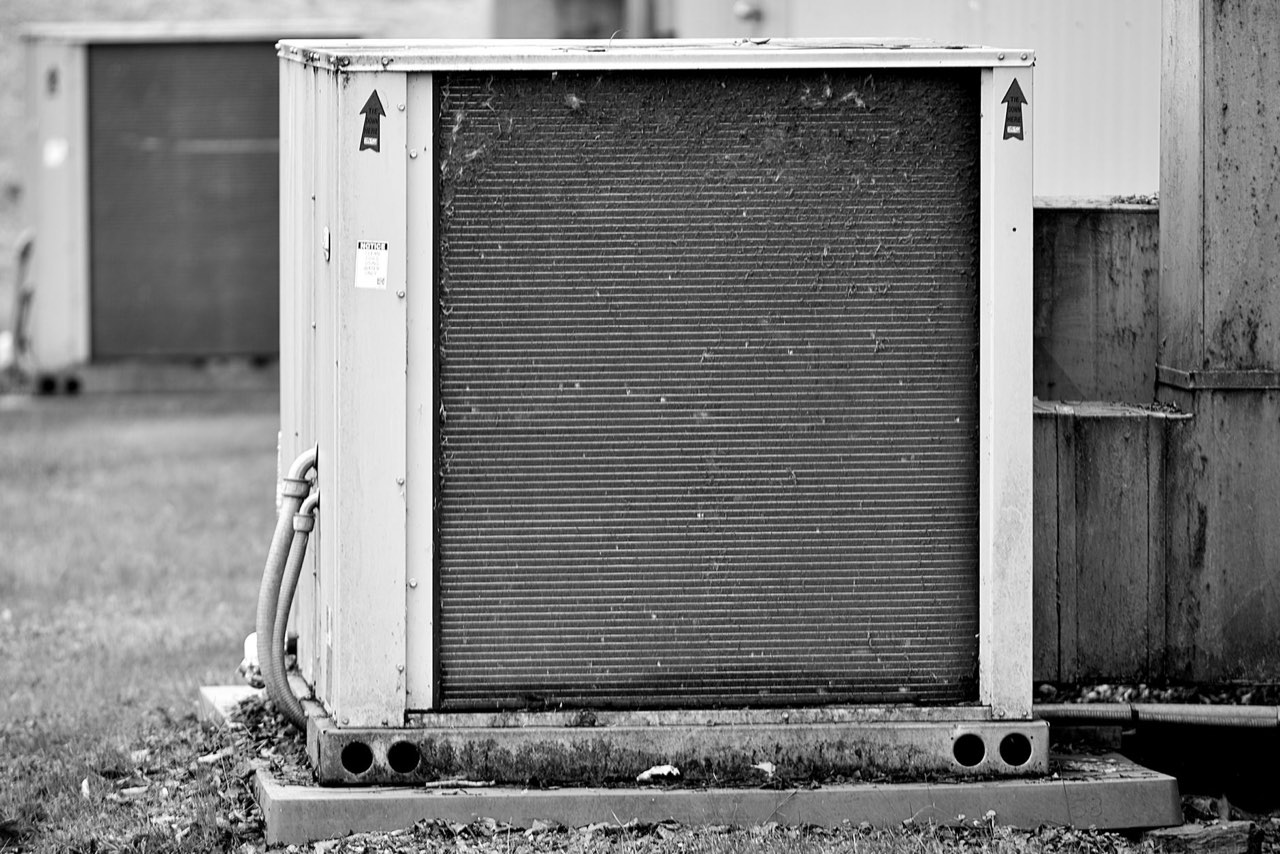 An outdoor condensing unit on a concrete pad, its fin coil dusty and streaked, in black and white