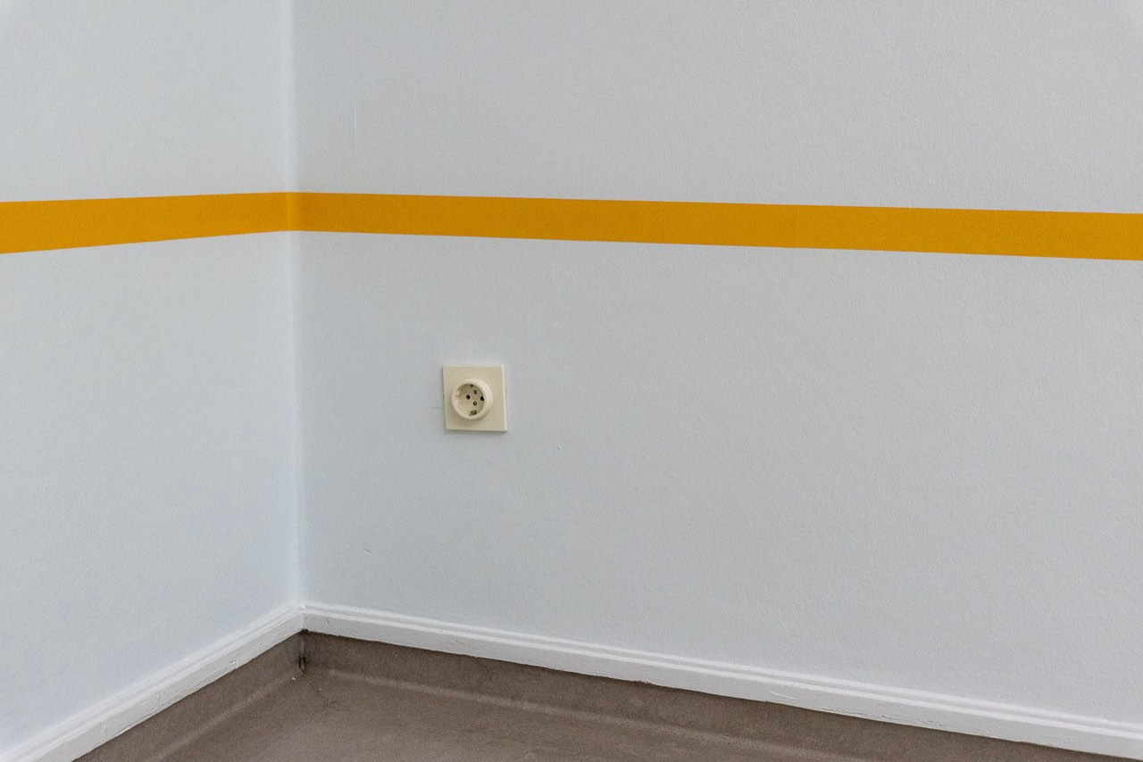 The corner of a pale painted room with a yellow band along both walls, a single socket and a white skirting board meeting a grey floor