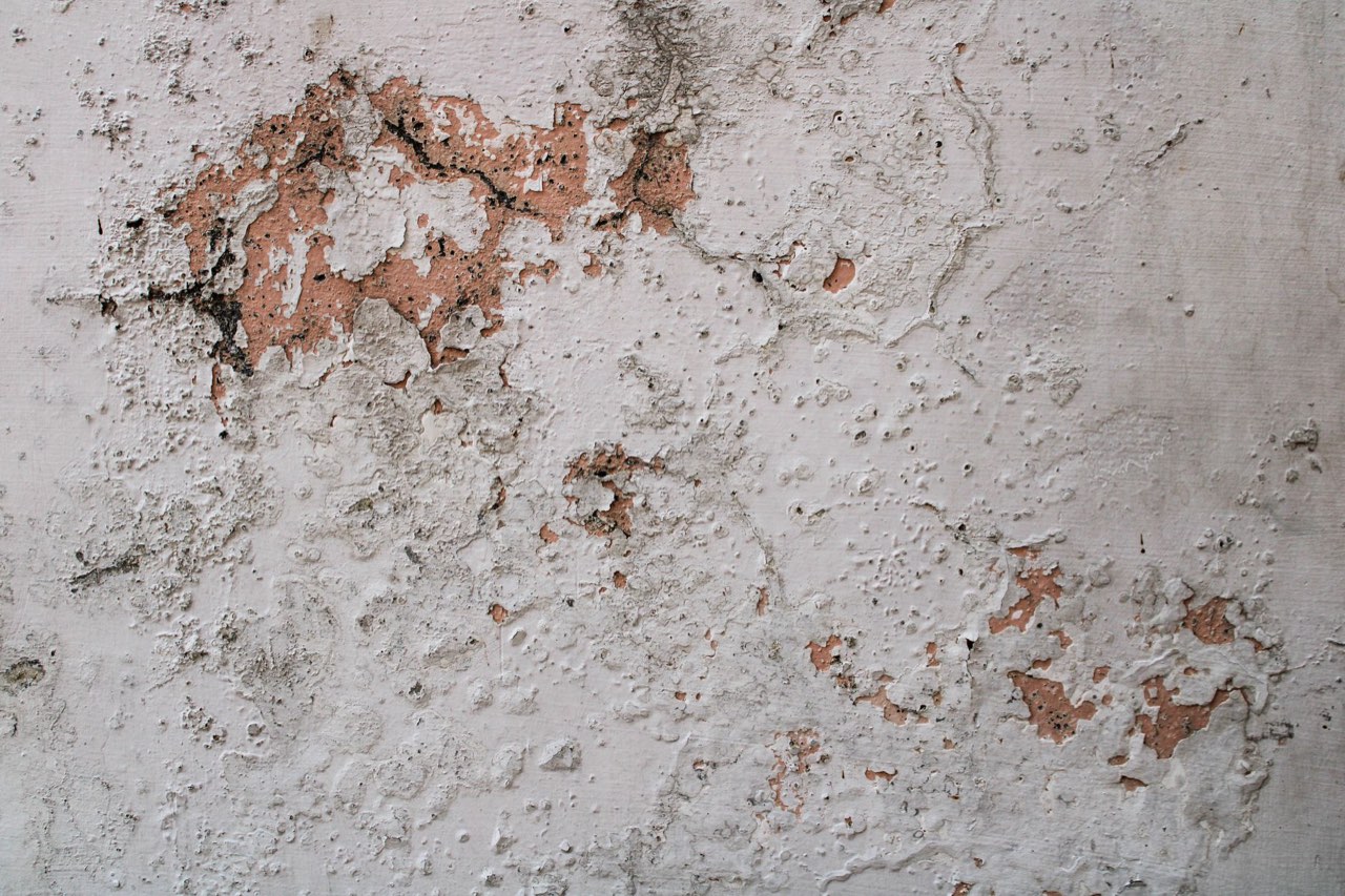A white painted wall blistered and flaking, with patches of bare pink plaster and dark staining