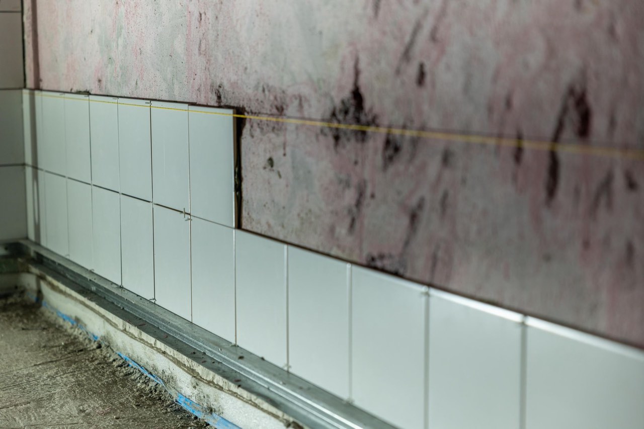 A wall half tiled in white up to a string line, bare render above it and spacer rails along the floor