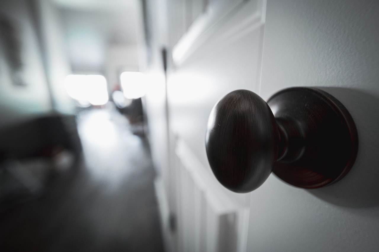 A dark bronze knob on a white panel door, close up, with an out of focus hallway behind it
